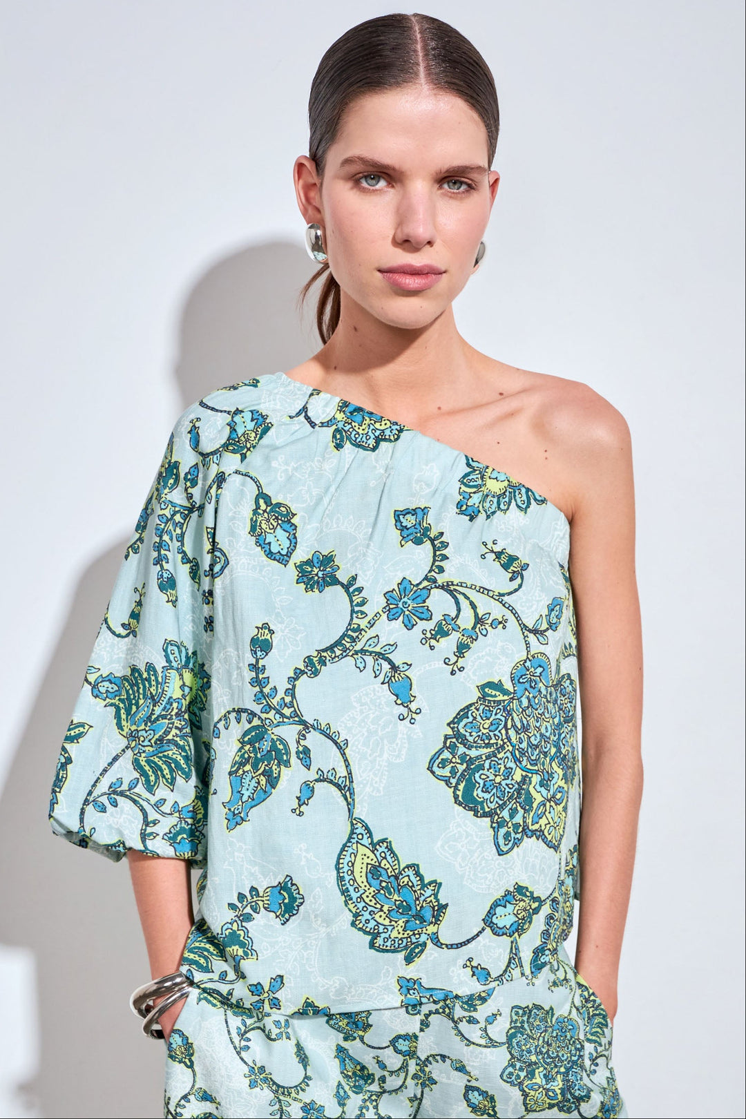 A woman modeling a one-shoulder light blue dress with a green and blue floral pattern, set against a plain light gray background.
