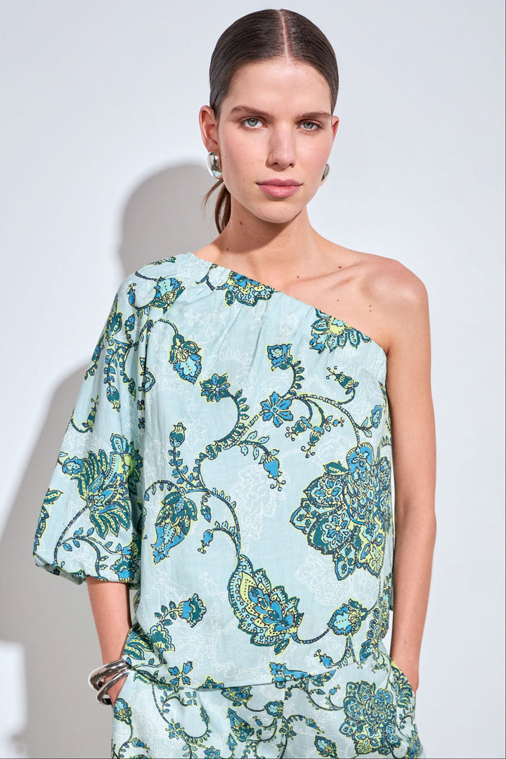 A woman modeling a one-shoulder light blue dress with a green and blue floral pattern, set against a plain light gray background.