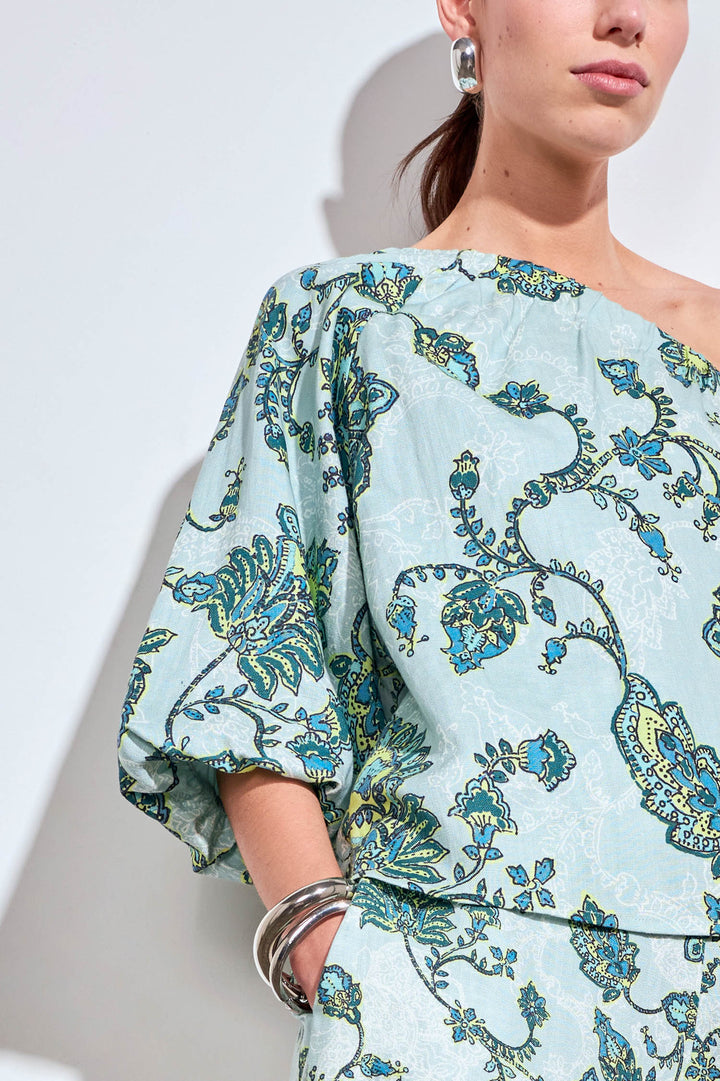 A close-up of a model wearing a light blue-green top with a blue and green paisley pattern, featuring voluminous elbow-length sleeves and a draped neckline.