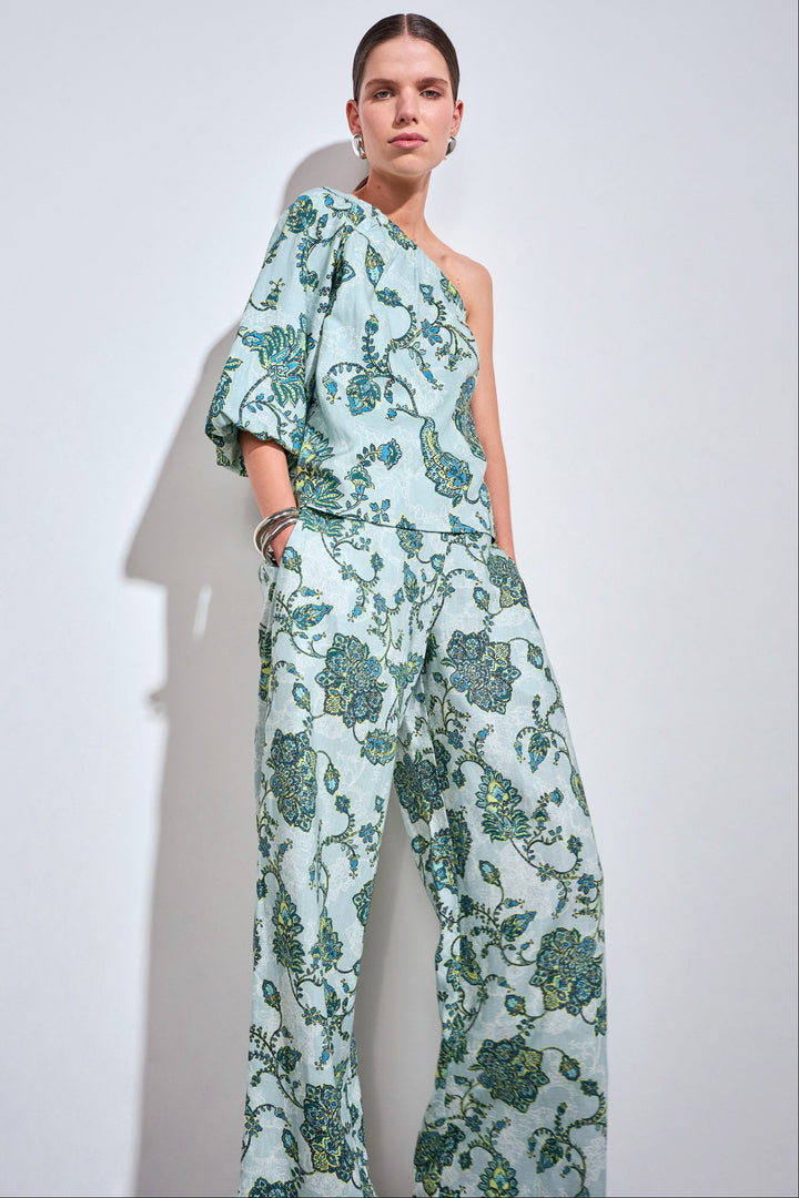 A model wearing a one-shoulder jumpsuit with a green and blue floral pattern on a light mint background, standing against a white wall with a soft shadow.