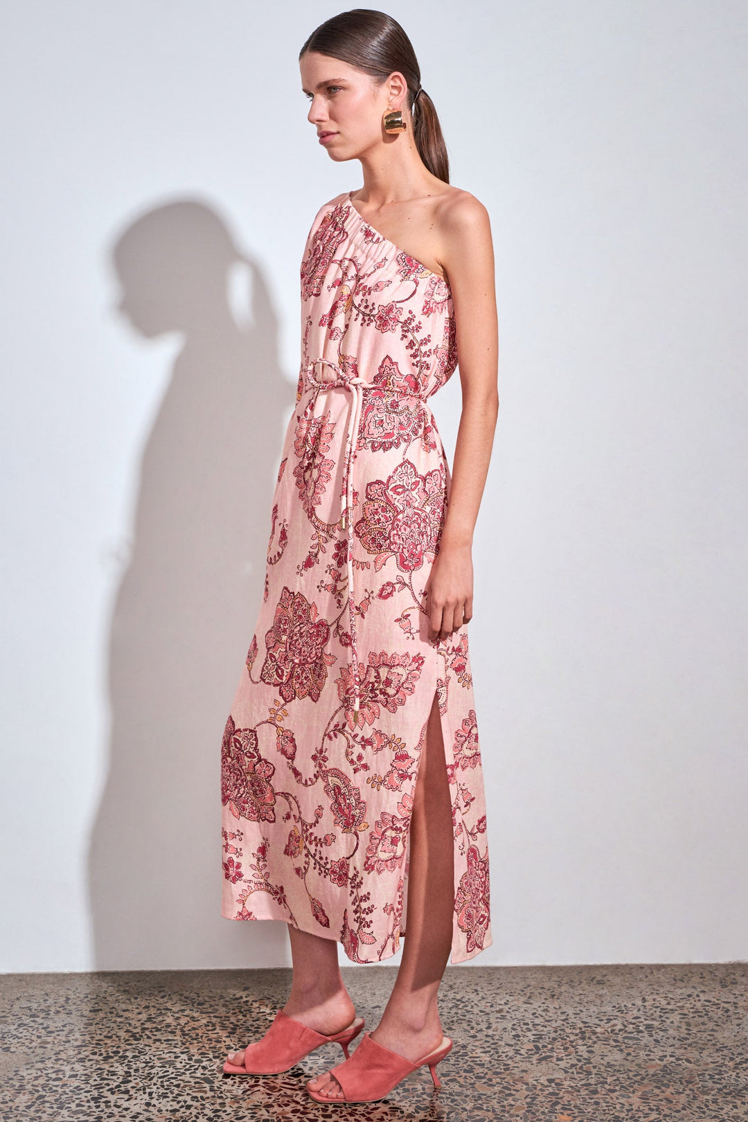 A woman models a one-shoulder midi dress with a pink and red botanical pattern, featuring a tie waist and a side slit. She is standing against a plain white wall with a strong shadow cast to the left.