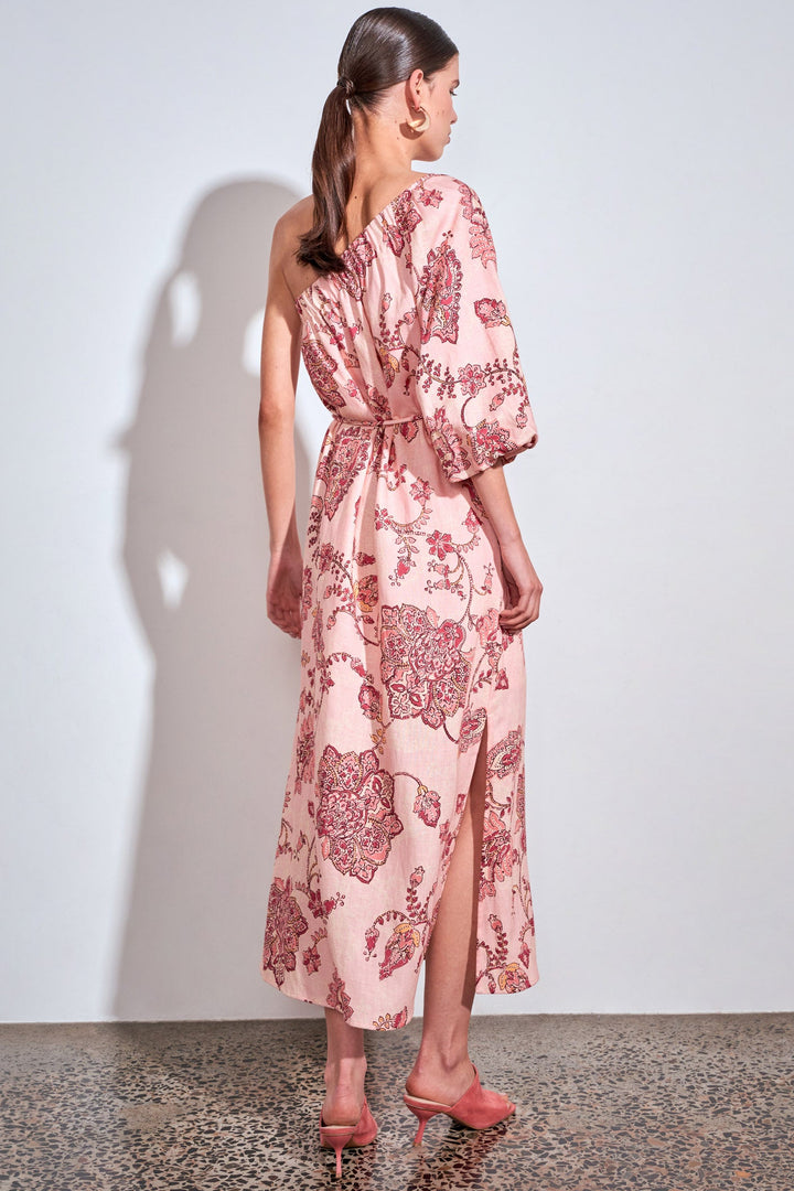 A woman stands with her back to the camera wearing a light pink one-shoulder midi dress with a dark red botanical pattern, paired with matching pink high heels against a white wall.
