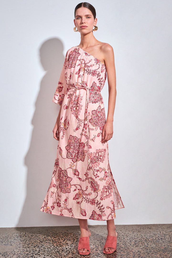 A model stands against a light gray wall wearing a one-shoulder midi dress with a pink and red floral pattern, paired with matching pink open-toe heels.