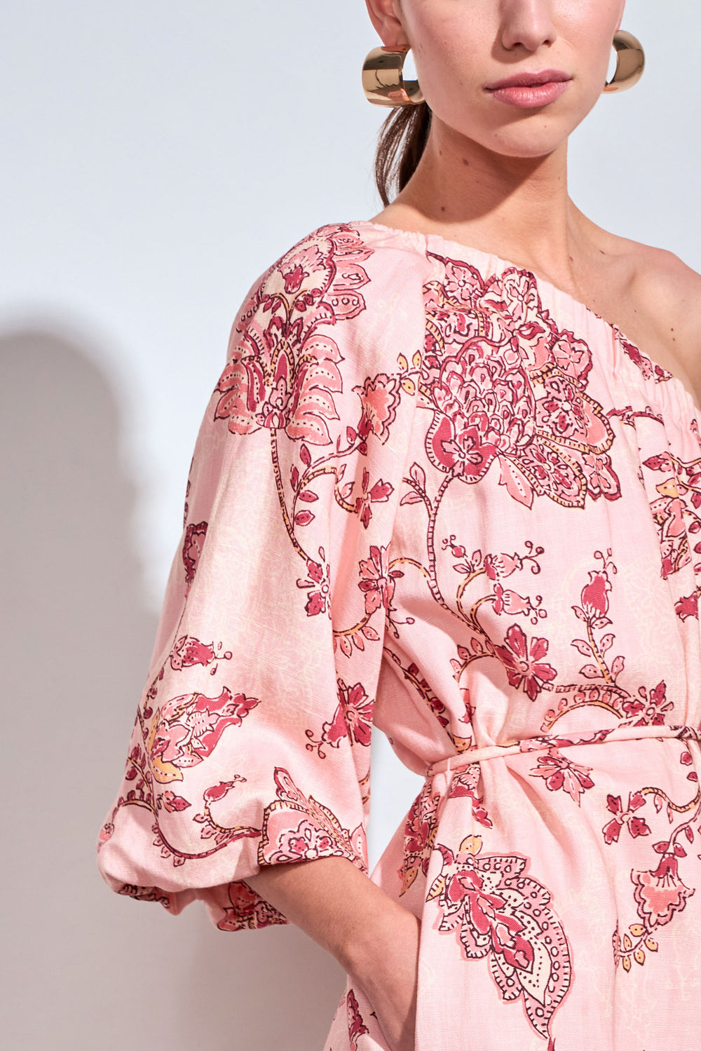 A close-up of a model wearing a light pink one-shoulder dress with a red and gold floral paisley print, featuring a tie waist and elbow-length sleeves.