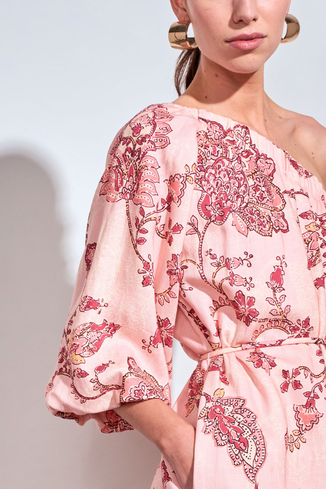 A close-up of a model wearing a light pink one-shoulder dress with a red and gold floral paisley print, featuring a tie waist and elbow-length sleeves.