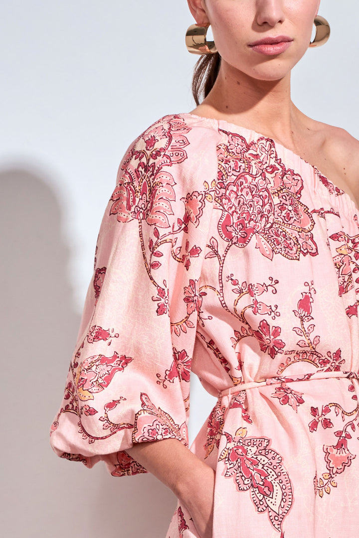 A close-up of a model wearing a light pink one-shoulder dress with a red and gold floral paisley print, featuring a tie waist and elbow-length sleeves.