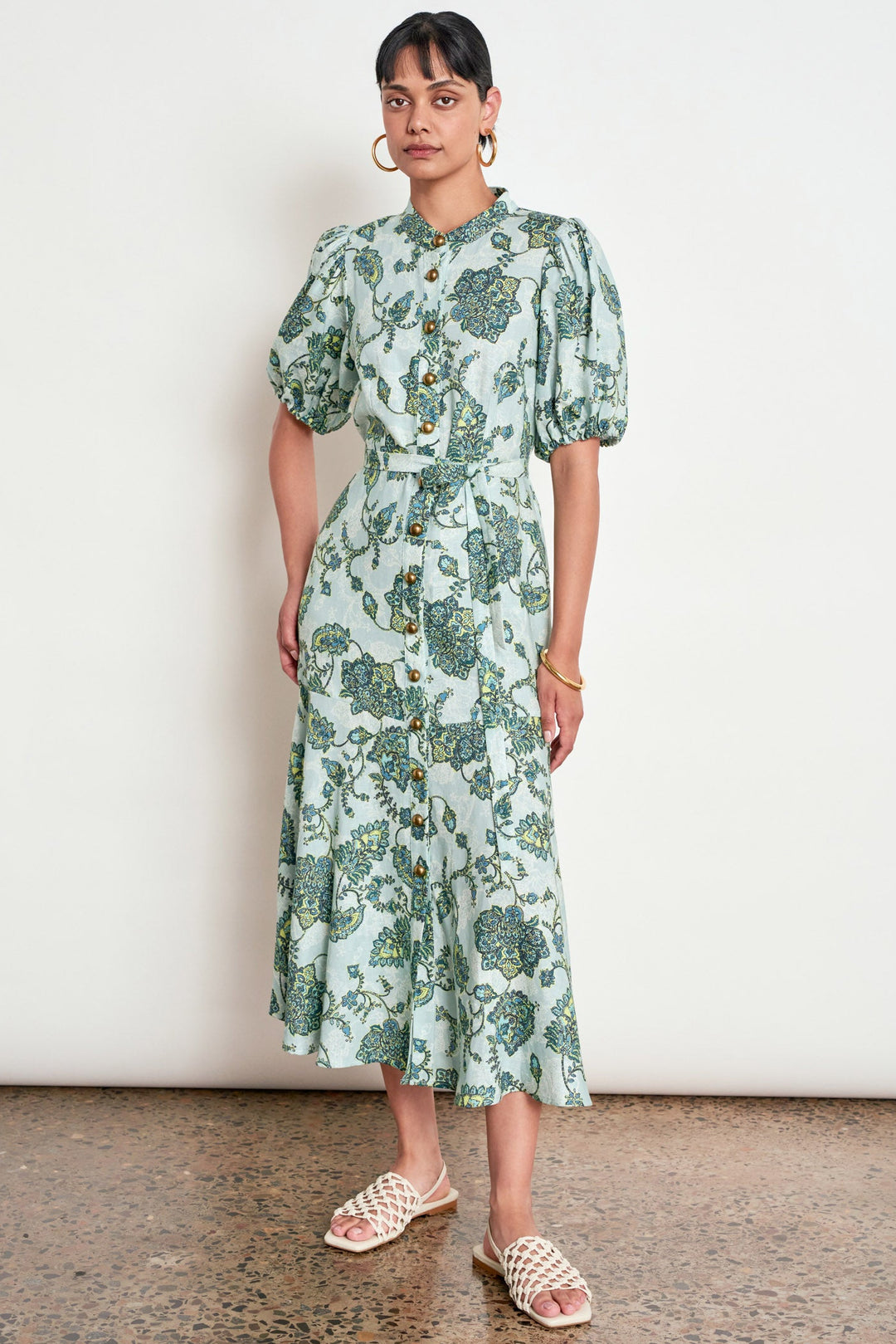 A woman modeling a light green midi dress with a blue and green floral print, featuring puff sleeves, a button-up front, and a tie-waist detail, paired with woven slide sandals against a plain white wall.