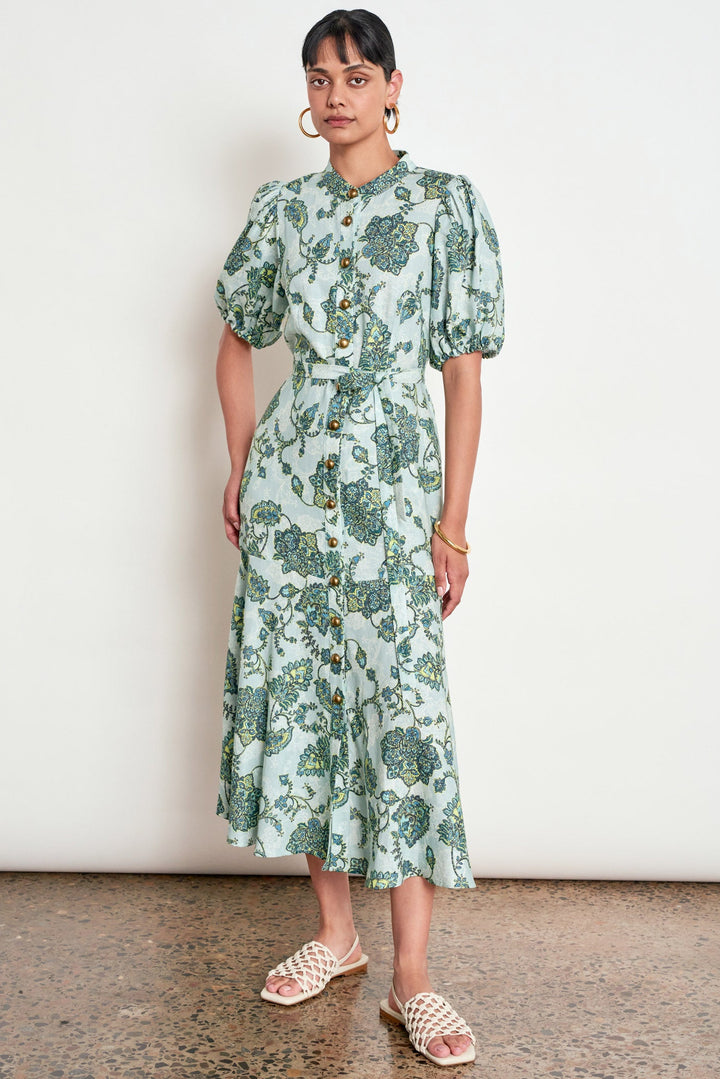 A woman modeling a light green midi dress with a blue and green floral print, featuring puff sleeves, a button-up front, and a tie-waist detail, paired with woven slide sandals against a plain white wall.