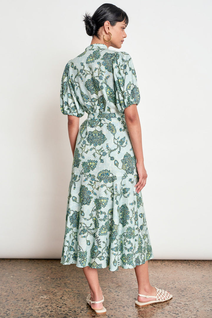 A woman is shown from the back wearing a light green midi dress with a dark green floral pattern, puff sleeves, and a gathered waist. She is standing on a speckled brown floor against a plain white wall.
