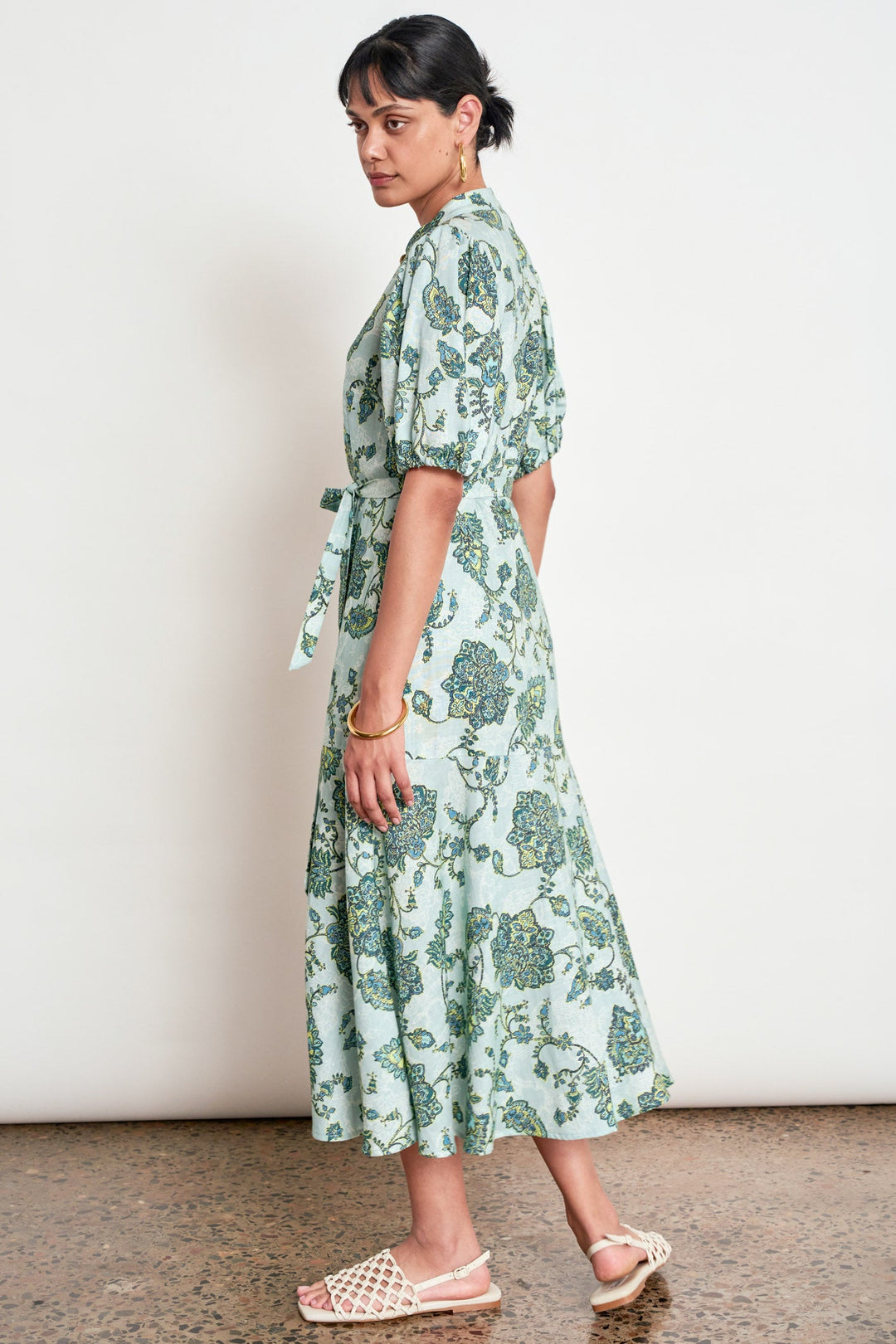 A woman modeling a light green and teal floral print midi dress with puff sleeves and a tie waist, paired with beige woven sandals, set against a plain white wall.