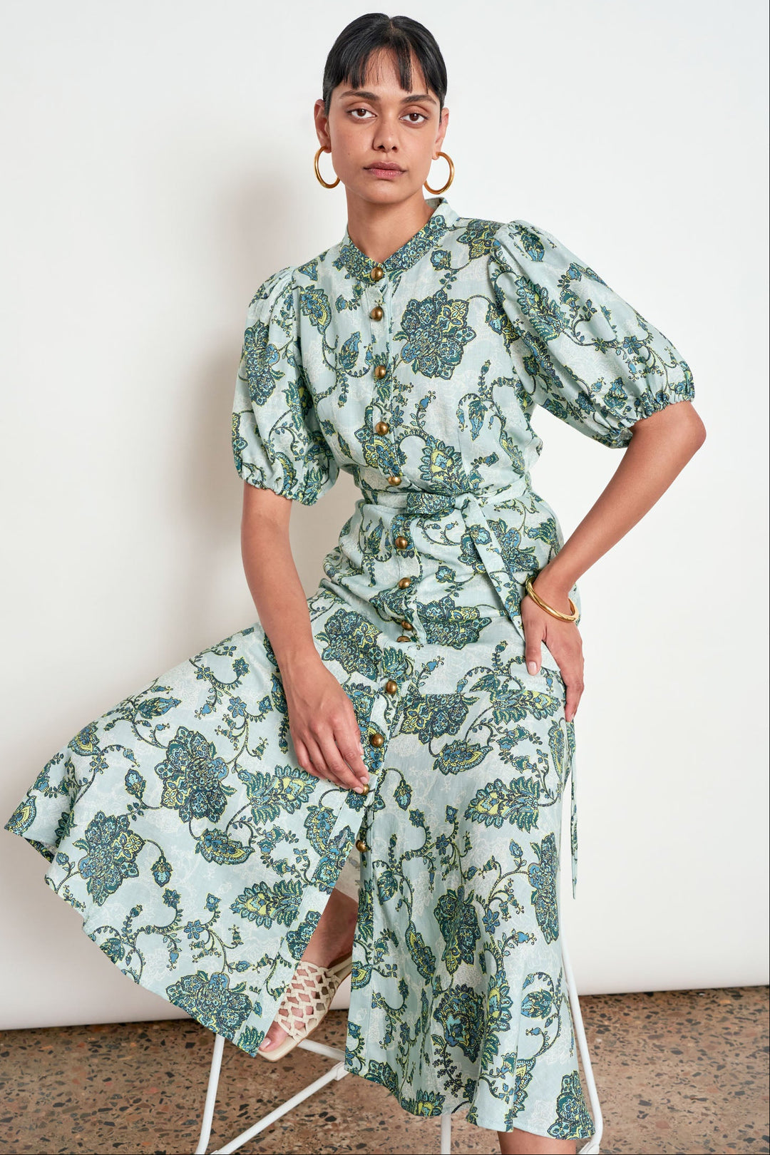 A woman models a light sage green midi dress with a blue-green botanical pattern, featuring puff sleeves, a button-up front, and a matching tie belt, while sitting on a white metal stool against a white wall.