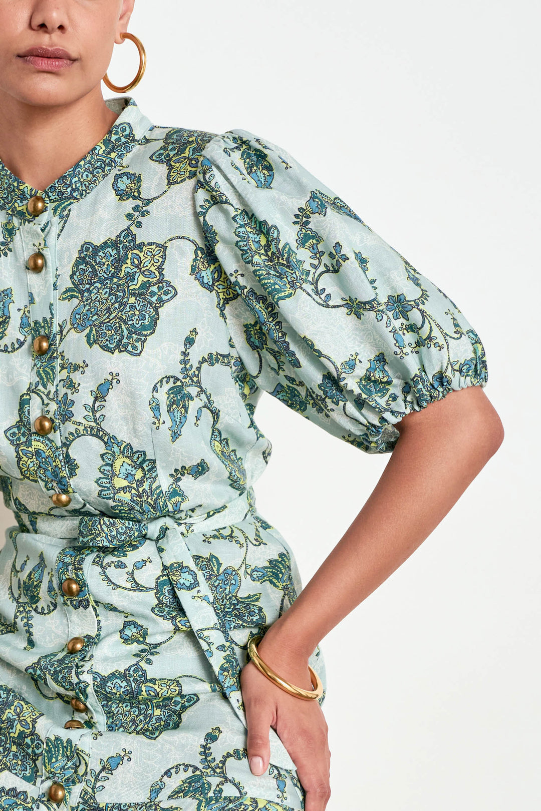 A close-up of a model wearing a light sage green dress with a blue and green floral pattern, featuring puff sleeves and gold-tone buttons.