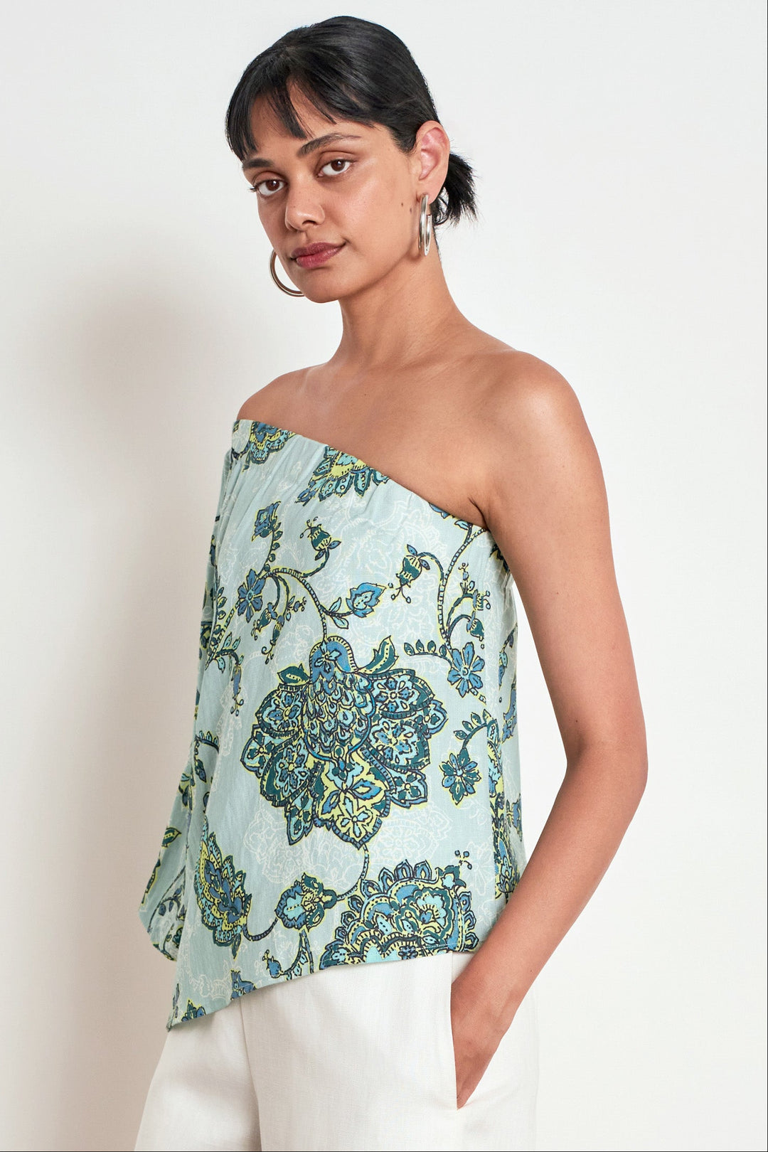 A woman modeling a strapless one-shoulder top with a light blue-green base and an intricate blue, green, and yellow floral pattern, paired with white pants against a plain off-white background.