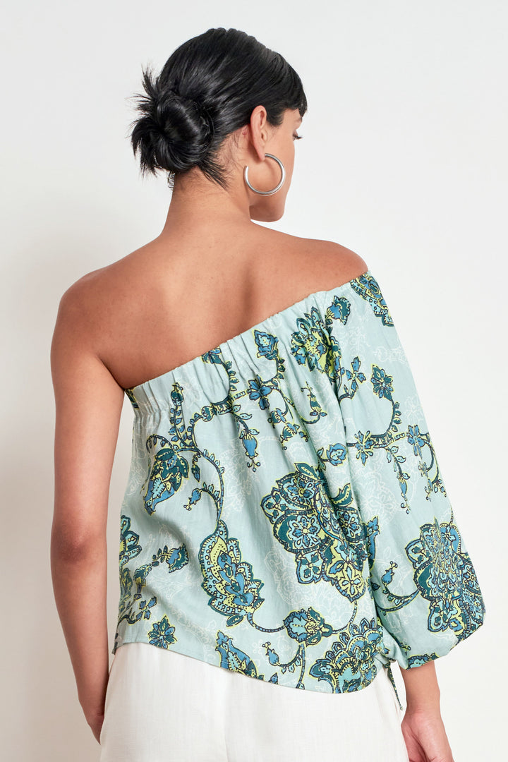 A woman shown from the back wearing a one-shoulder off-the-shoulder top with a light blue-green base and a blue and yellow floral pattern, paired with white bottoms against a plain white background.