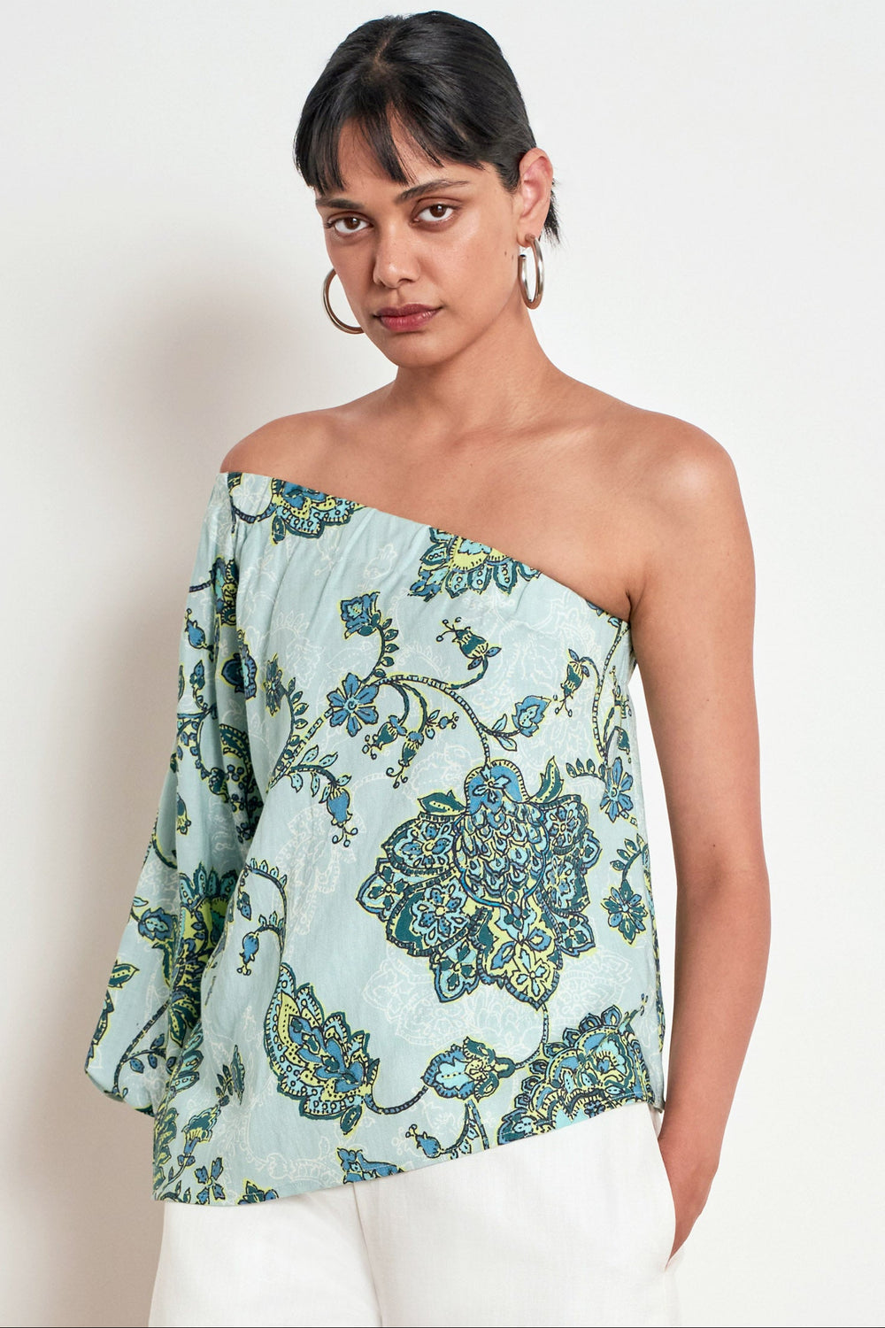 A woman modeling a one-shoulder strapless top with a light blue-green base and a blue, green, and yellow floral pattern, paired with white pants against a plain off-white background.