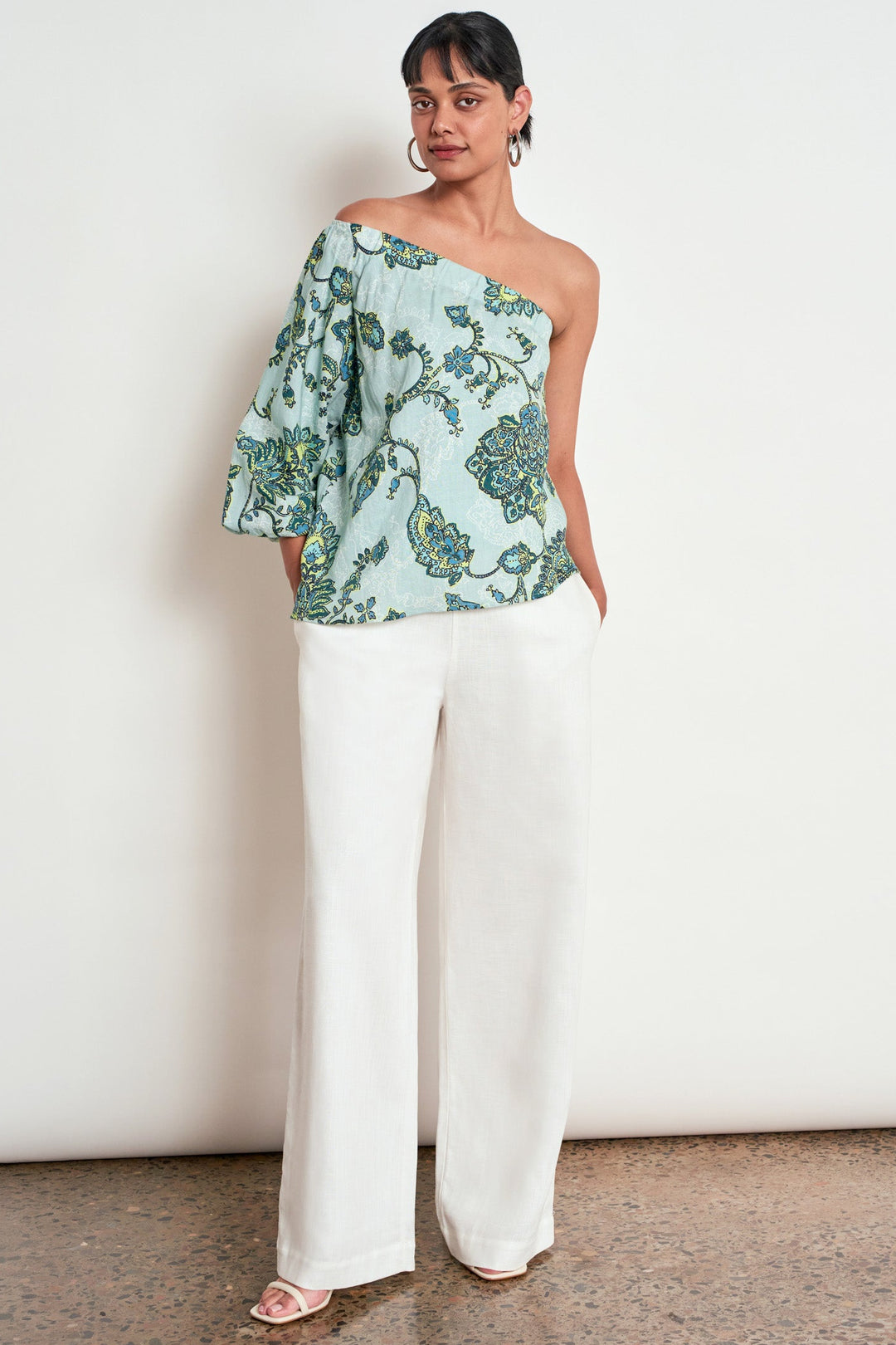 A woman modeling a one-shoulder top with a light blue-green floral print and white wide-leg pants, standing against a plain off-white wall.
