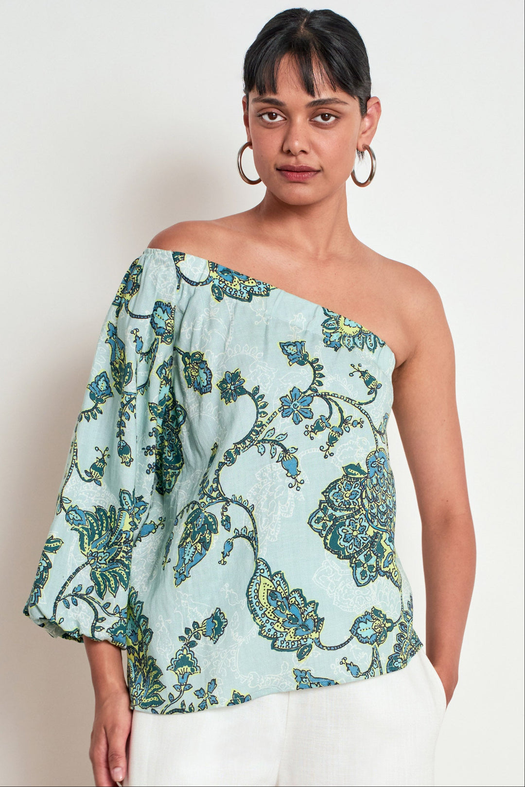 A woman modeling a light blue off-the-shoulder blouse with a green and blue floral paisley pattern, paired with white pants, against a plain off-white background.