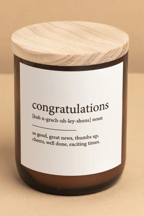 A brown candle jar with a white label that reads 'congratulations' and definitions, topped with a wooden lid.