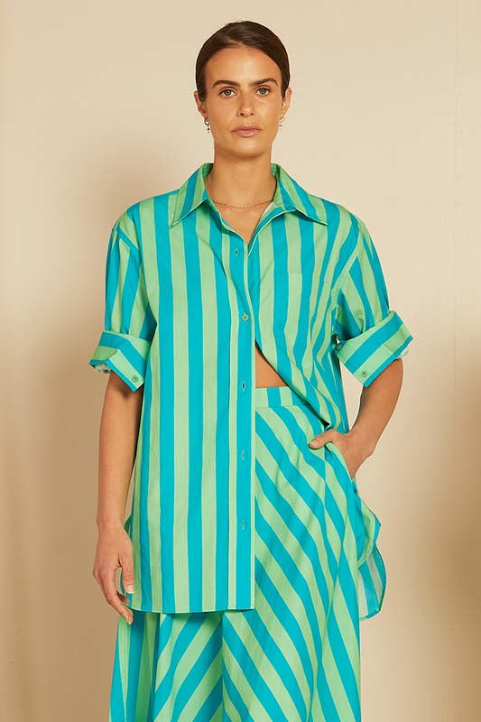 A model wearing a striped shirt in shades of blue and green with a relaxed fit and rolled-up sleeves.
