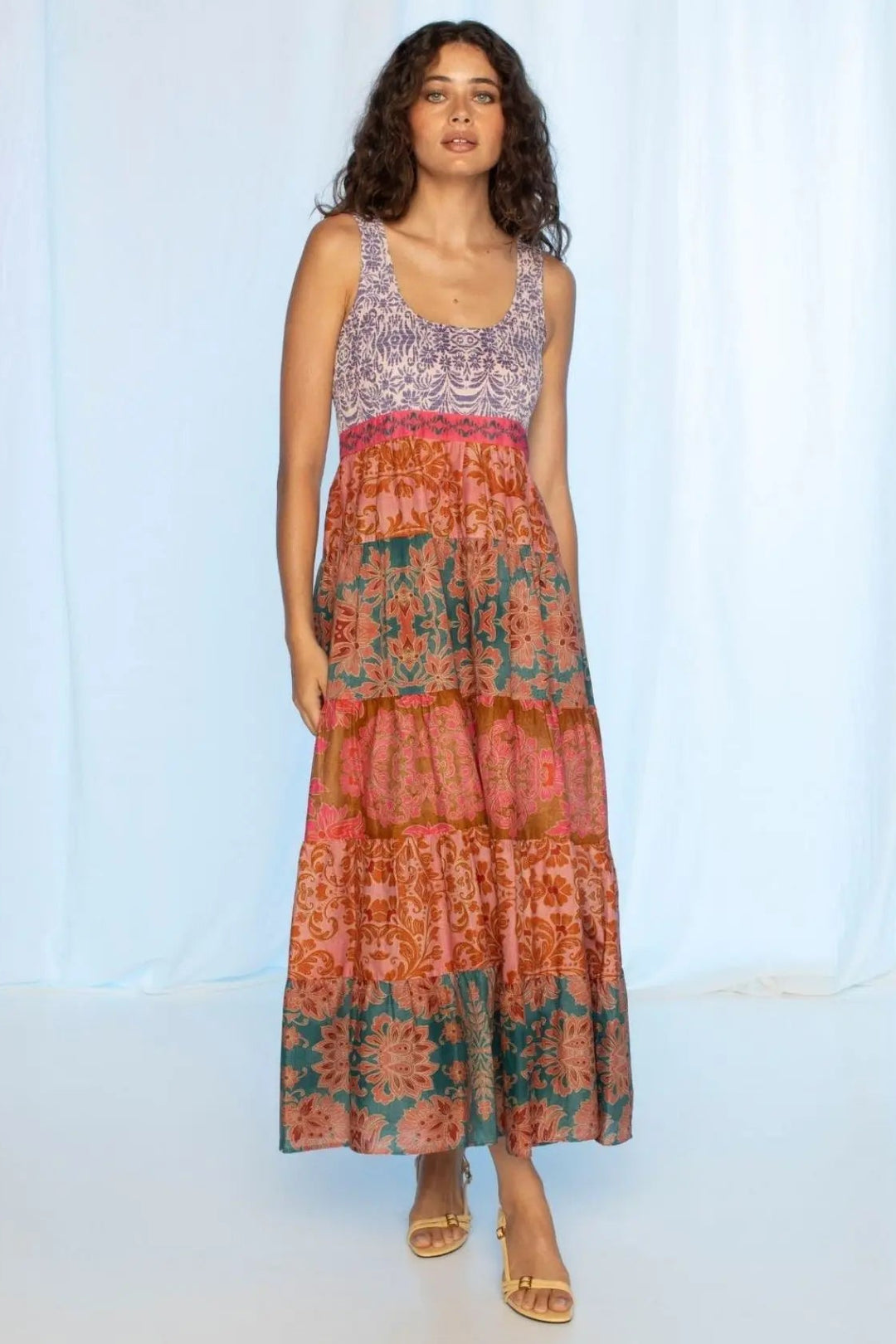 Woman wearing a colorful dress with a patterned top and tie-dye skirt against a light blue curtain background.