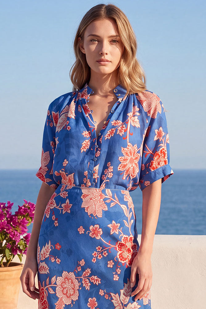 A woman stands outdoors by the sea wearing a blue midi dress with a coral and pink floral print. The setting includes a white wall, blue ocean, and potted pink flowers.