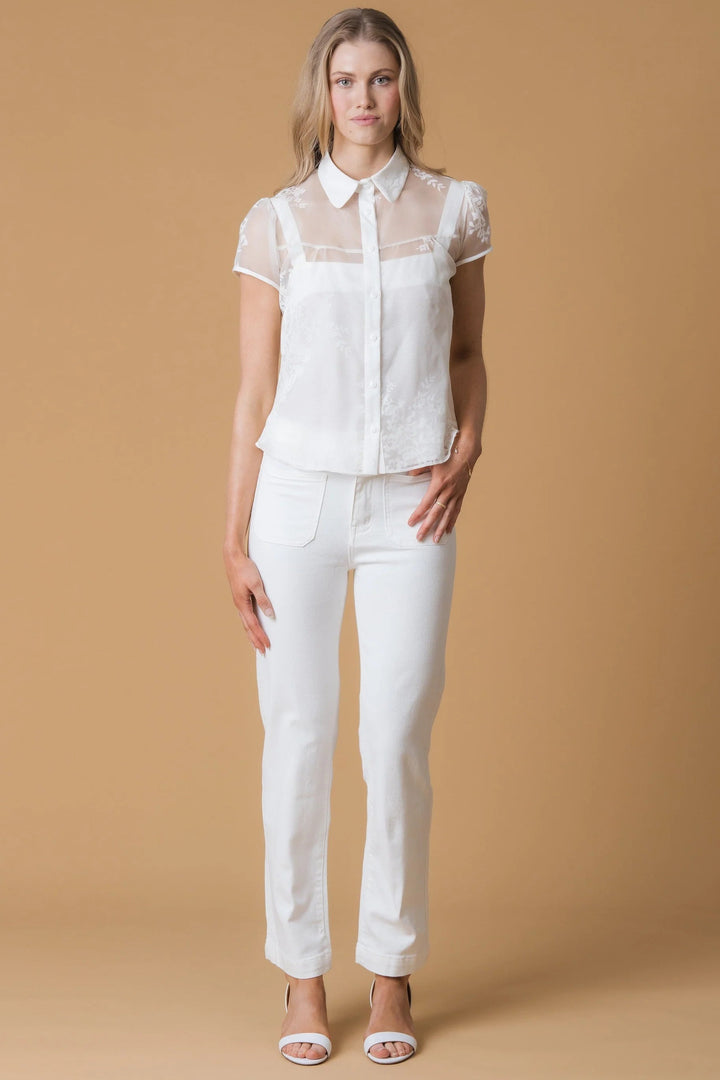 A woman modeling a white short-sleeve blouse with sheer mesh sleeves and white pants, standing against a solid tan studio background.