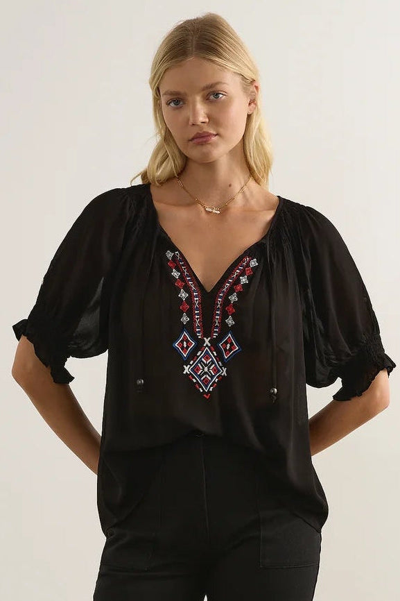 A blonde model wearing a black sheer puff-sleeve blouse with a V-neckline and colorful geometric embroidered detailing in red, blue, and white, paired with black pants against a light gray studio background.