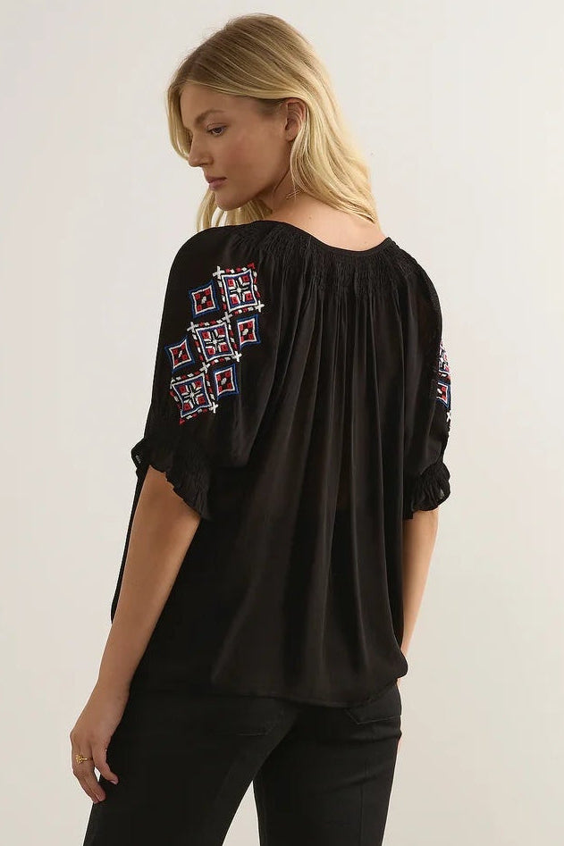 A woman wearing a black short-sleeve blouse with colorful geometric embroidery on the upper sleeves, paired with black pants, photographed against a plain light background.