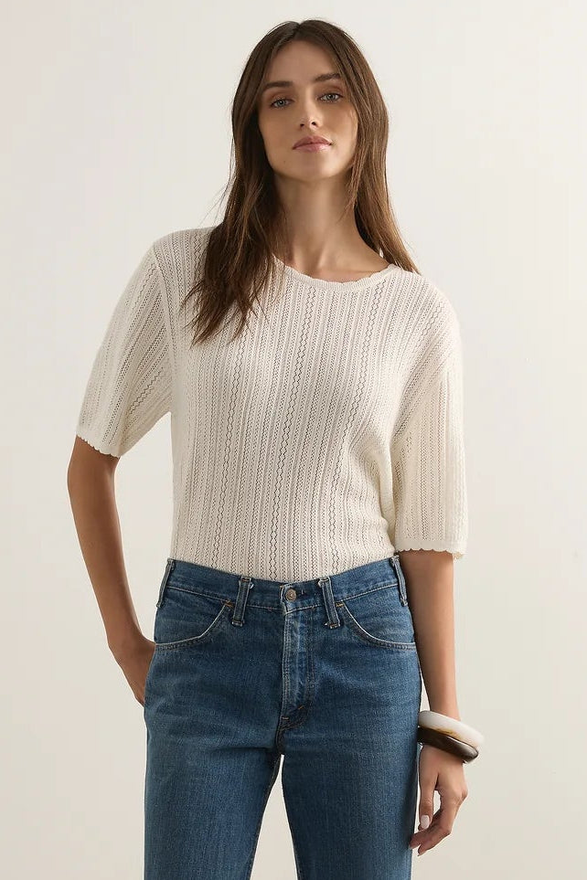 A woman wearing a cream-colored, short-sleeved ribbed knit top and blue jeans, standing against a plain off-white studio background.