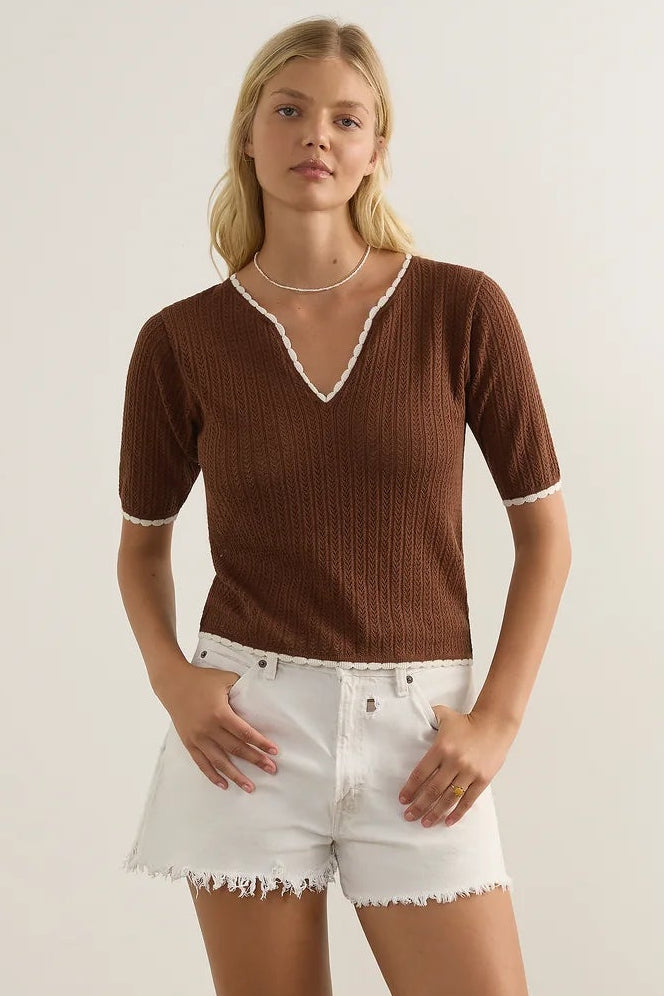 A woman modeling a brown ribbed short-sleeve V-neck top with white trim, paired with white frayed denim shorts and brown strappy sandals, standing against a plain off-white studio wall.