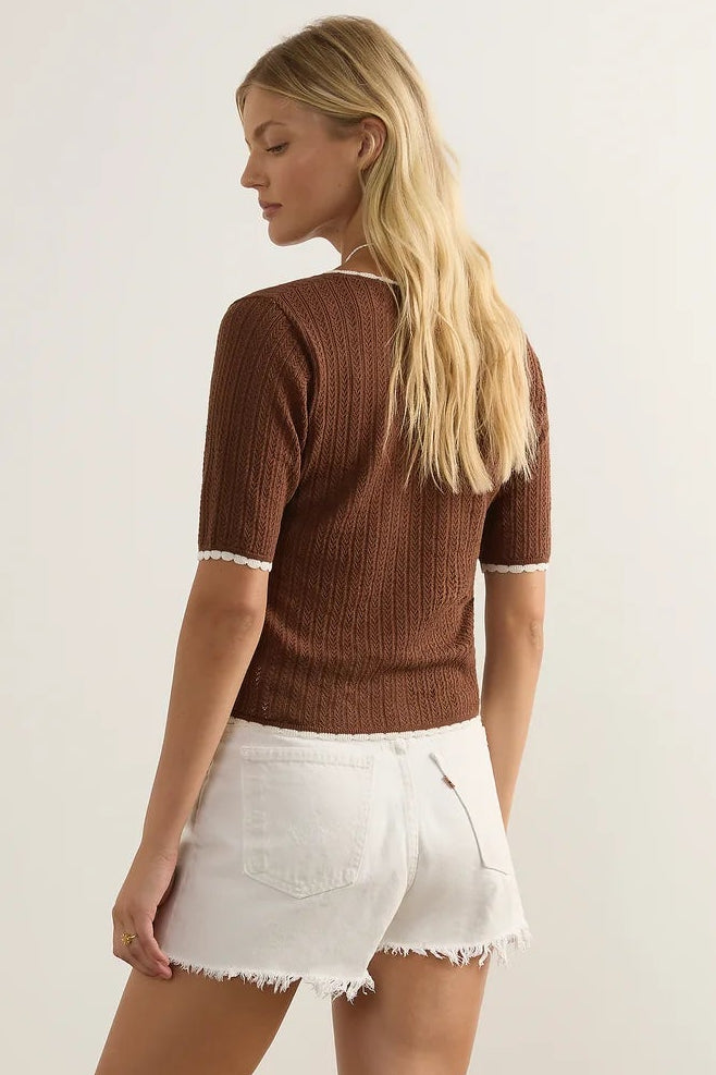 A woman is shown from the back wearing a brown short-sleeve ribbed knit top with white frayed denim shorts and brown strappy sandals, standing against a plain light beige studio background.