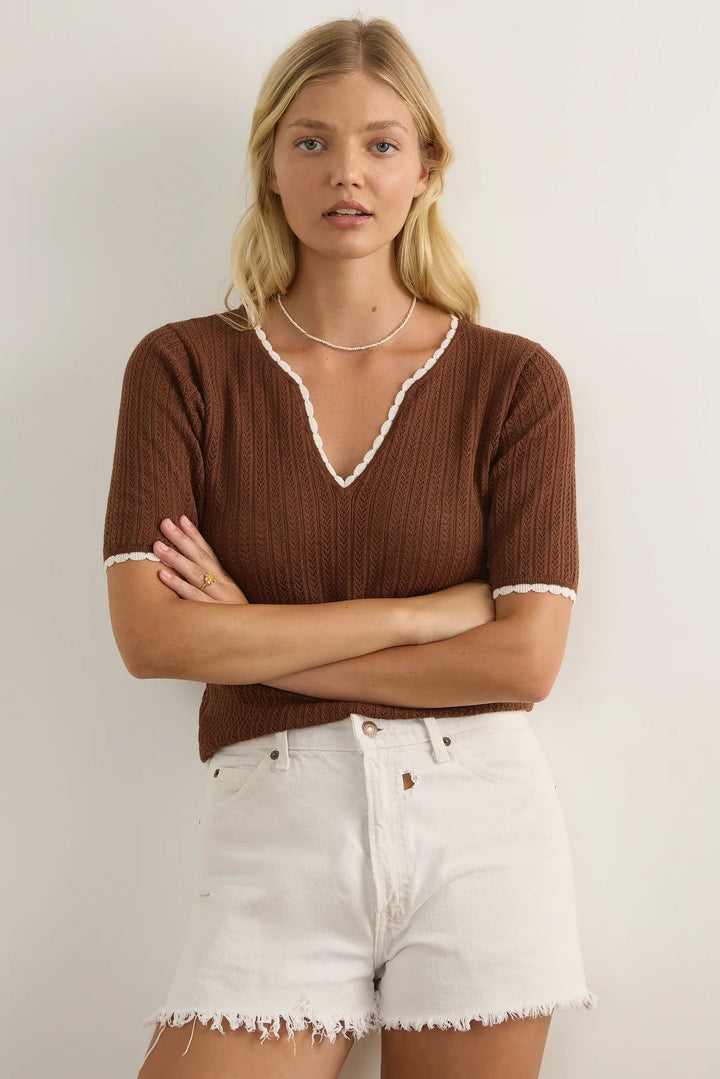 A blonde woman wearing a brown short-sleeve ribbed knit top with white lace trim at the V-neck and sleeve cuffs, paired with white denim shorts, standing against a plain off-white wall.