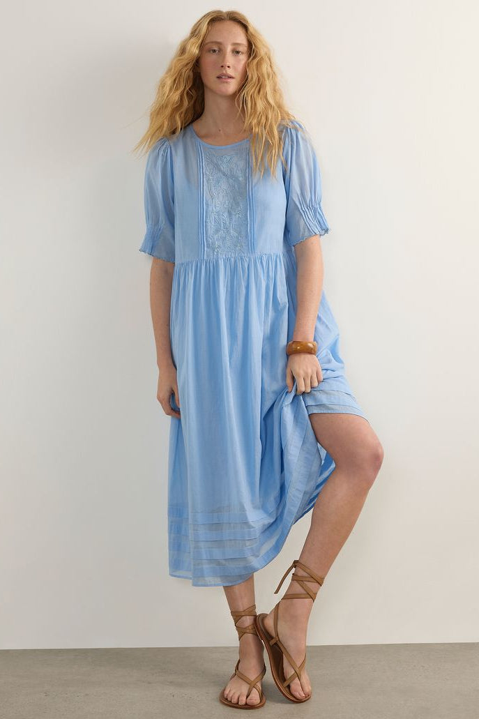 A woman modeling a light blue midi dress with short puff sleeves, a round neckline, and embroidered detailing on the chest, paired with brown strappy sandals against a plain off-white wall.