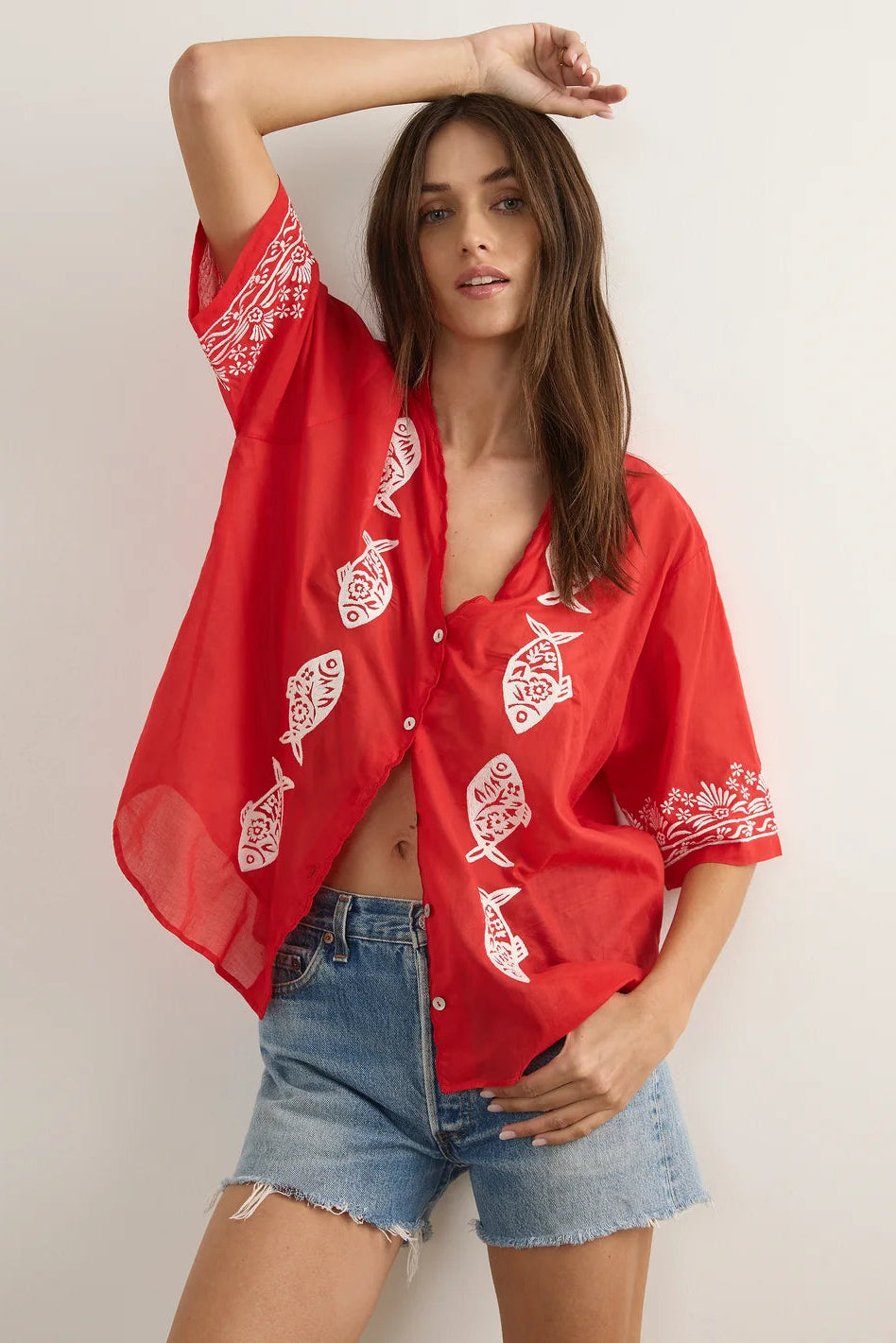 A woman modeling a red short-sleeve button-up shirt with white fish and tribal-style embroidery, paired with denim shorts, against a plain light background.