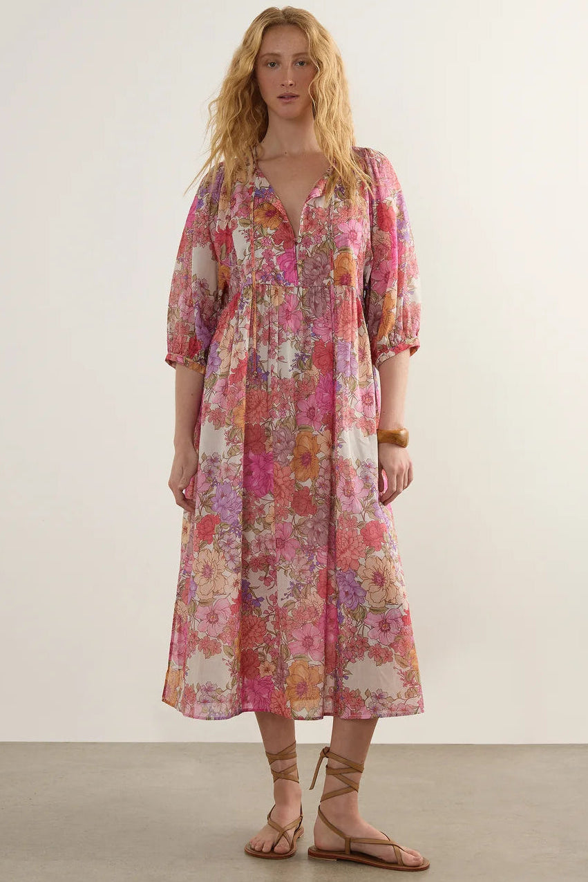 A woman modeling a long-sleeved midi dress with a vibrant pink, orange, and beige floral pattern, standing against a plain off-white studio wall.