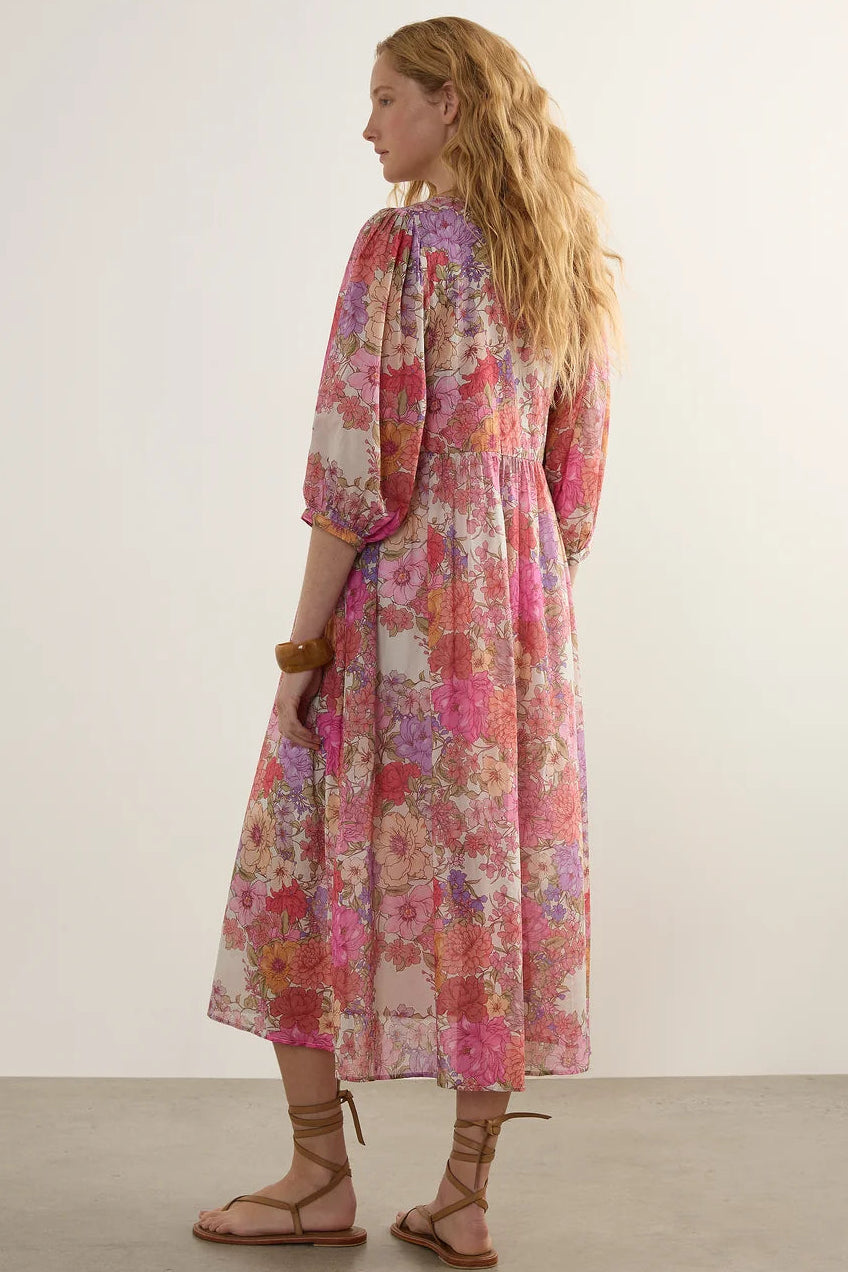 A woman modeling a long-sleeved midi dress with a vibrant pink, purple, and orange floral pattern, shown from a side-back angle against a neutral studio background.