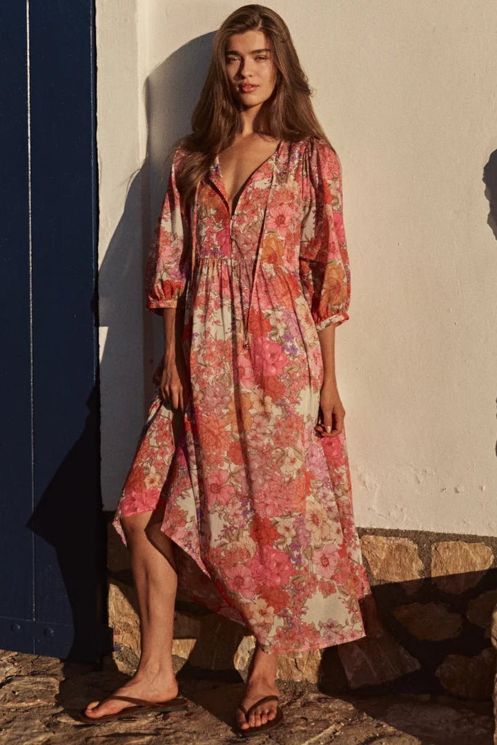A woman stands barefoot outdoors against a white wall and stone steps, wearing a long floral midi dress with three-quarter sleeves and a V-neck. The dress has a pink, orange, and cream floral pattern.