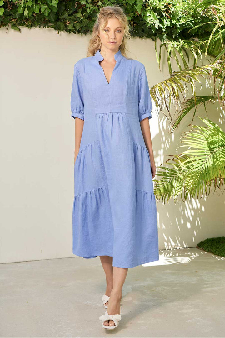A woman standing in a garden wearing a blue knee-length dress with short sleeves and a V-neckline.