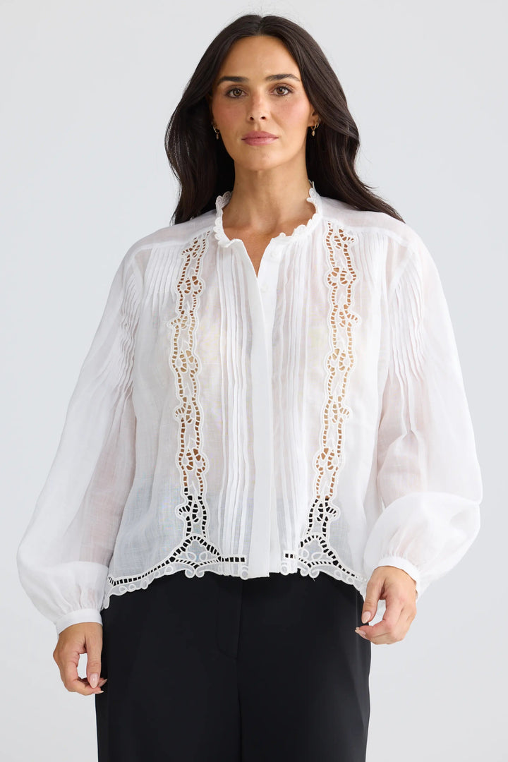 A woman wearing a white long-sleeve blouse with vertical lace applique panels down the front, paired with black trousers, photographed against a light gray studio background.