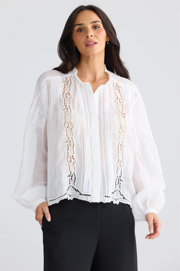 A woman modeling a white long-sleeve blouse with intricate gold and black embroidered lace details down the front, paired with black trousers, against a light gray studio background.