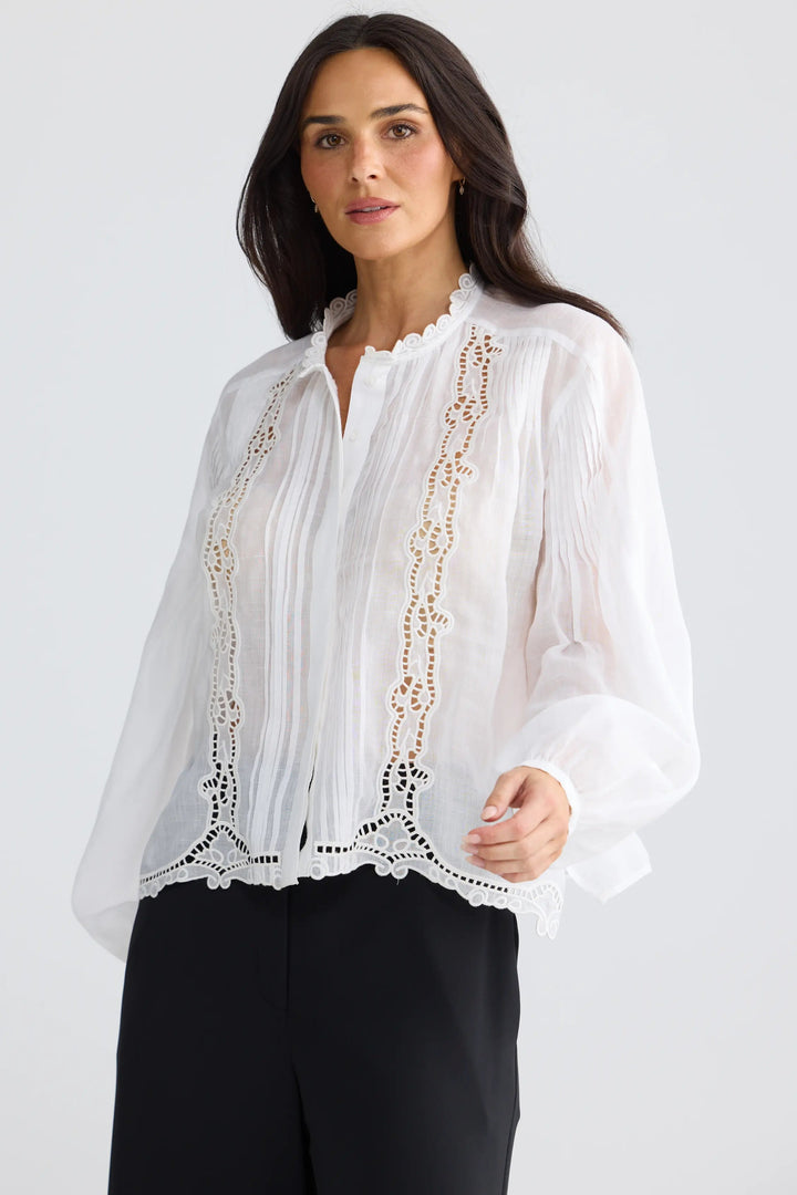 A woman modeling a white long-sleeve blouse with delicate lace and crochet embroidery on the front, paired with black trousers, against a light gray studio background.