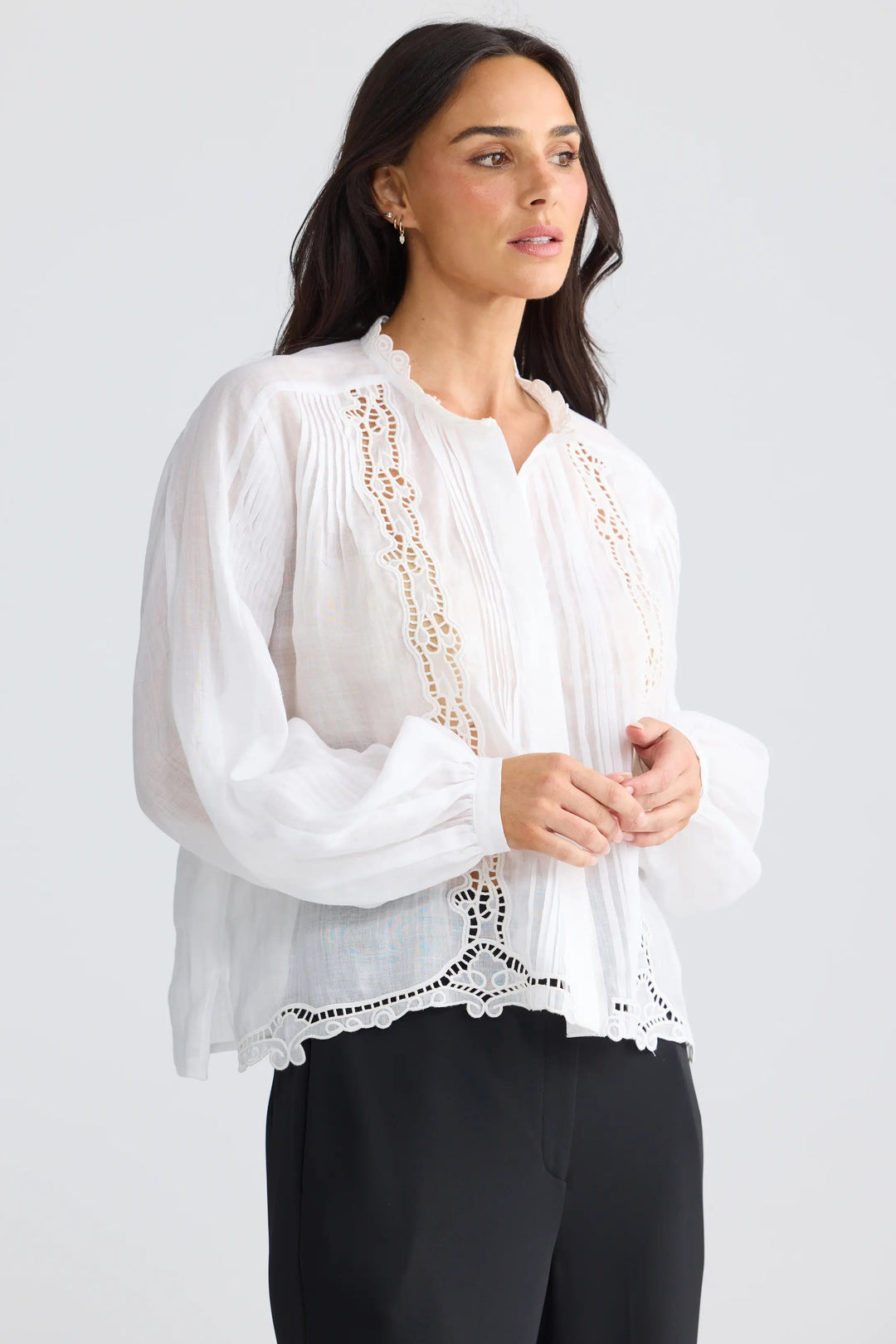 A woman modeling a white long-sleeve blouse with delicate lace embroidery and pleated detailing, paired with black trousers, against a light gray studio background.