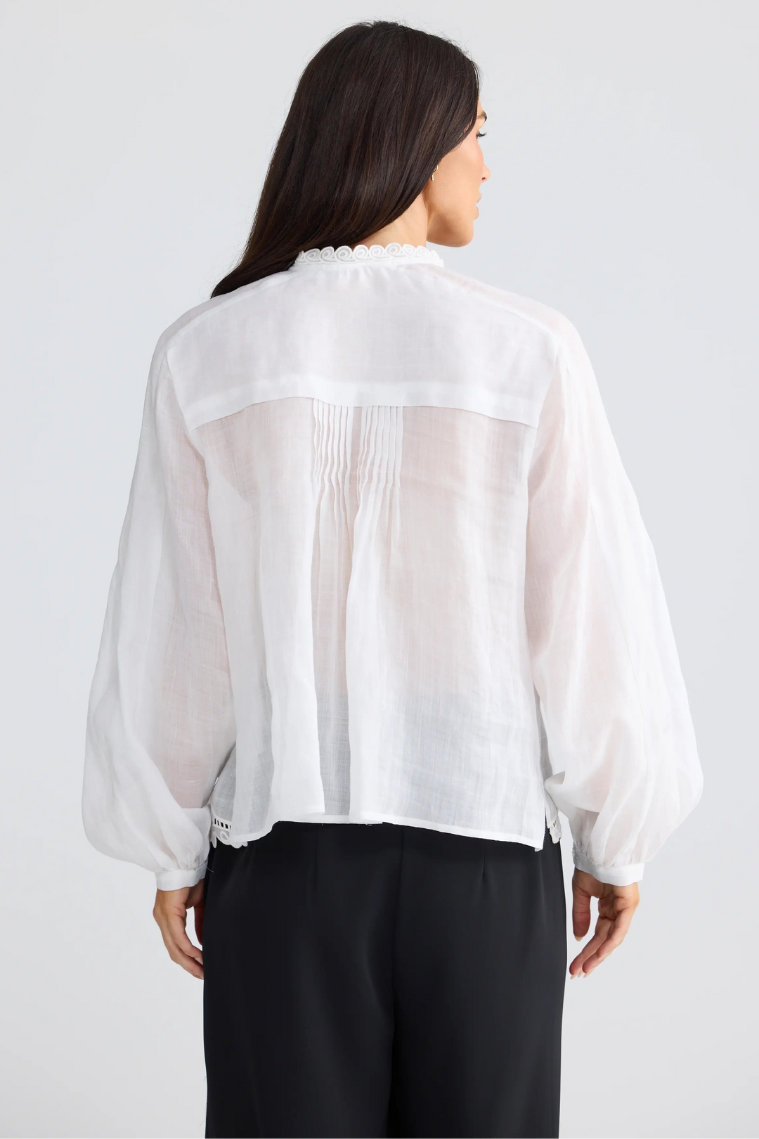 A woman viewed from the back wearing a white sheer long-sleeve blouse with gathered pleating at the back and black pants, against a light gray studio background.