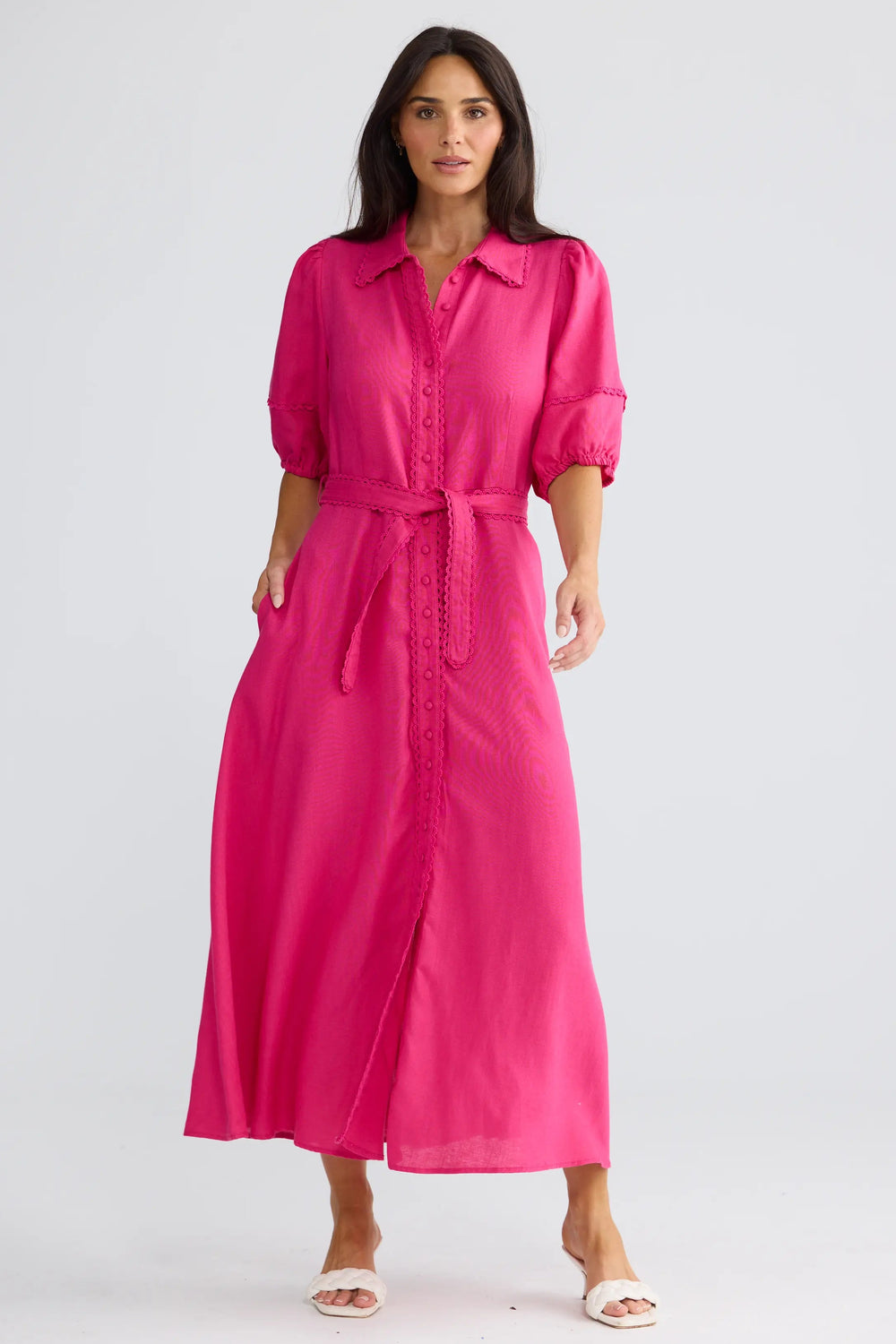 A woman modeling a bright pink midi shirt dress with three-quarter sleeves, a matching tie belt, and side pockets, paired with white slide sandals against a light gray studio background.