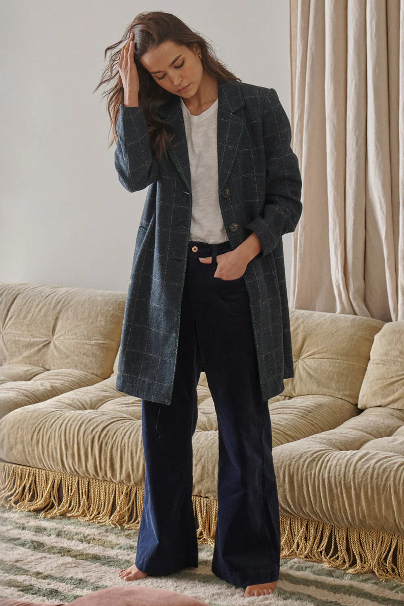 A person standing in a room with elegant decor wearing a navy coat with a windowpane check pattern and green contrast stitching, paired with dark trousers.