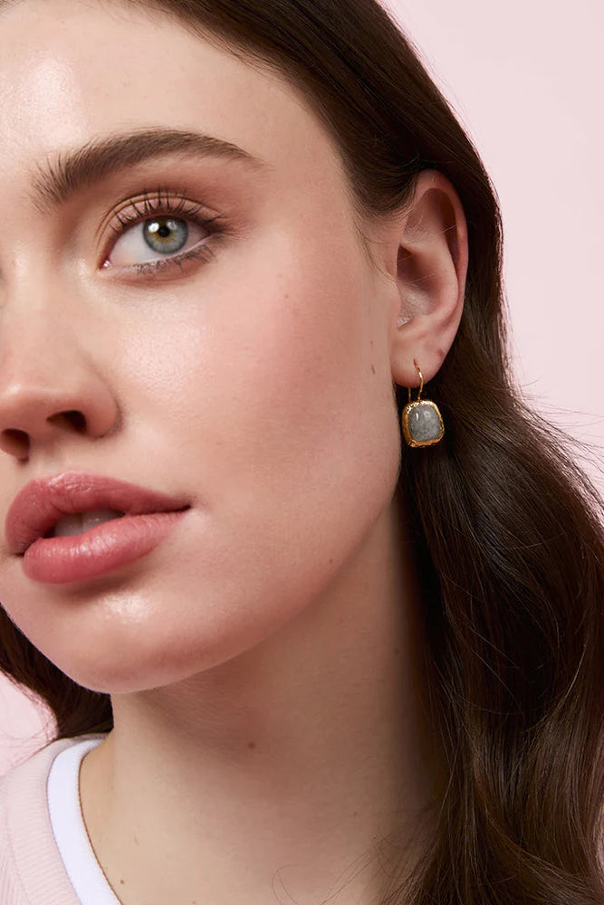 A close-up of a woman wearing a small gold dangle earring with a square, iridescent blue-green stone, set against a solid pale pink background.