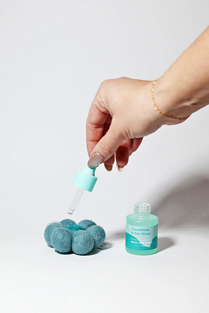 Hand holding a fragranced oil dropper over flower shaped felt air freshener, next to a bottle of the fragranced oil on a light gray background.