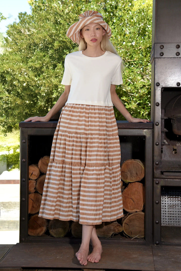 A woman standing by a firewood storage area, wearing a coffee stripe patterned tiered skirt with a t-shirt style bodice, posing with one foot raised on a log.