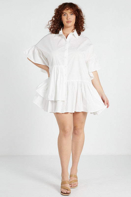 A female model wearing a white mini dress with a classic collared neckline, half sleeve with ruffle at cuff, and asymmetrical gathered skirt tier at the front. The model is standing against a neutral background and is posing in a relaxed stance.