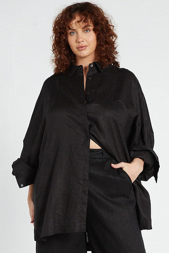 A model wearing a black oversized linen shirt with sleeves scrunched and a curved hem, paired with matching pants.